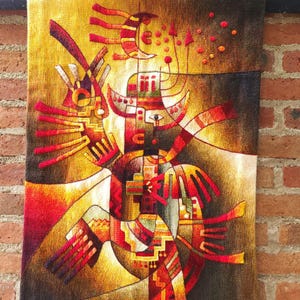 May include: A woven tapestry with an abstract design in shades of gold, red, and brown. The artwork features geometric shapes and stylized figures, with a central motif resembling a bird or mythical creature. The tapestry is rectangular and hangs against a brick wall.