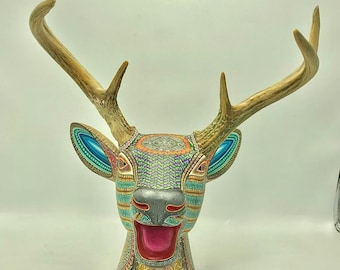 May include: A colorful wooden deer head sculpture with intricate patterns and a bright pink open mouth. The antlers are made of natural wood and are attached to the head.