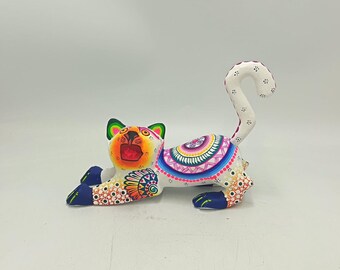 May include: A colorful, hand-painted wooden cat figurine in a reclining pose. The cat features a white base with intricate patterns in orange, blue, pink, and green. The tail is curved, and the cat's face is open-mouthed.