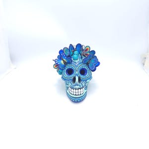 May include: A decorative blue skull figurine with intricate patterns and a butterfly crown. The skull features detailed designs and white teeth. The butterflies are in shades of blue and have colorful accents.