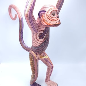 May include: A hand-carved wooden monkey figurine with intricate details and vibrant colors. The monkey is painted in shades of pink, purple, orange, and yellow, with a gold accent on its tail. The figurine is standing on its hind legs with its arms raised above its head.