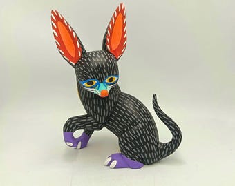 May include: A hand-carved, whimsical black fox figurine with a textured pattern. The fox features large orange and white ears, blue and yellow facial details, and purple paws. This folk art piece is a unique decorative item.
