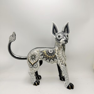 May include: A standing Xoloitzcuintli dog figurine featuring intricate black, white, and gold hand-painted patterns. The sculpture displays detailed geometric sunburst motifs, swirling filigree, and dotted textures across its entire body.