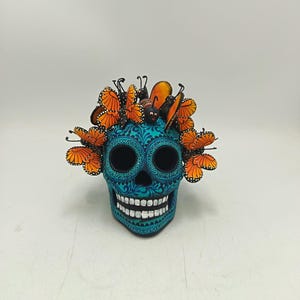 Könnte beinhalten: Dekorativer Totenkopf in Türkis und Blau mit floralem Muster. Der Totenkopf hat große Augenhöhlen und ein breites Grinsen mit weißen Zähnen. Orangefarbene und schwarze Schmetterlingsdekorationen schmücken den oberen Teil des Schädels.