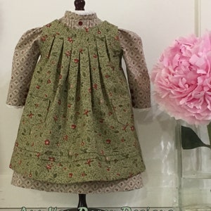 Pode incluir: Um vestido de estampa floral verde e marrom com saia franzida e dois bolsos. O vestido est&aacute; em um manequim e tem uma gola de renda branca.