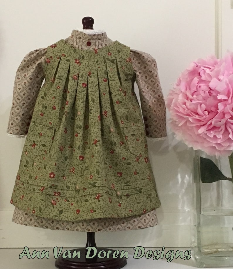 Pode incluir: Um vestido de estampa floral verde e marrom com saia franzida e dois bolsos. O vestido est&aacute; em um manequim e tem uma gola de renda branca.