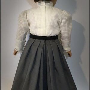 May include: A doll wearing a white blouse with a black velvet belt and a gray skirt. The doll has brown hair styled in a bun.