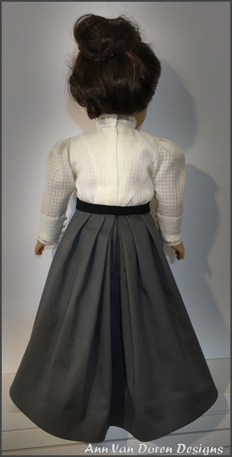 May include: A doll wearing a white blouse with a black velvet belt and a gray skirt. The doll has brown hair styled in a bun.