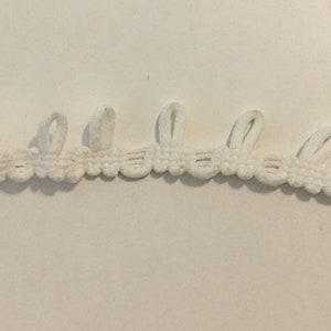 May include: A length of white decorative trim with a scalloped edge and looped accents. The trim is made of a woven material and is suitable for various craft and sewing projects. The trim is curved and lies on a white surface.