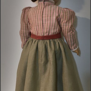 May include: A doll wearing a pink and green striped blouse with a red belt and a long brown skirt.