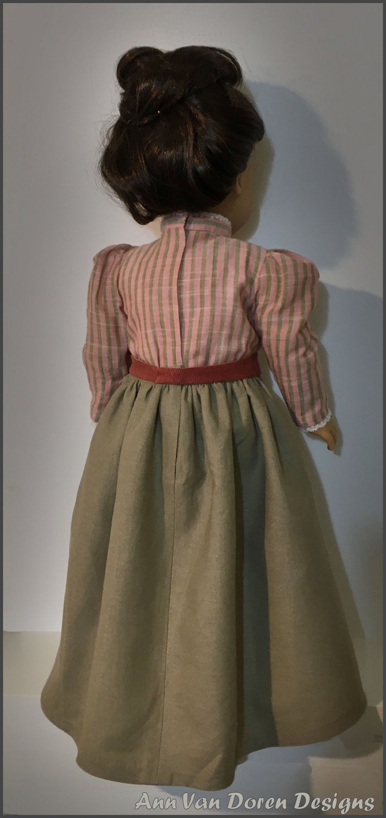 May include: A doll wearing a pink and green striped blouse with a red belt and a long brown skirt.