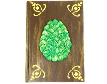 May include: A brown wooden journal with a green dragon egg design on the front cover. The egg is made of a textured material and has a shiny finish.
