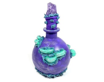 Könnte beinhalten: Eine violette Glasflasche mit einem dekorativen Stopfen, der mit einem violetten Kristall verziert ist. Die Flasche ist mit türkisfarbenen und grünen Akzenten verziert.