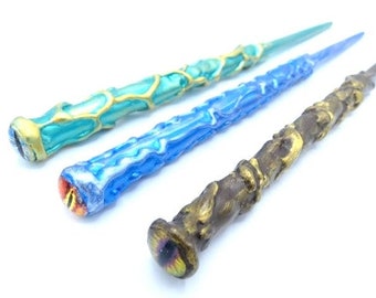 Puede incluir: Tres varitas decorativas con diferentes acabados de color. La primera varita es de color verde azulado con detalles dorados, la segunda es azul con detalles plateados y la tercera es marrón con detalles dorados.