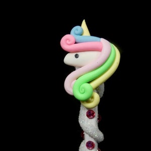 May include: A white unicorn head with a rainbow mane and a pink horn. The unicorn head is attached to a white stick with pink glitter.