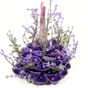 May include: A purple and pink seashell centerpiece with a tall, slender, pink base and a cluster of purple seashells. The centerpiece is decorated with small purple flowers and beads.