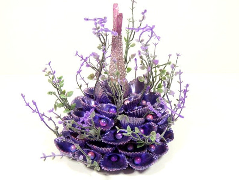 May include: A purple and pink seashell centerpiece with a tall, slender, pink base and a cluster of purple seashells. The centerpiece is decorated with small purple flowers and beads.
