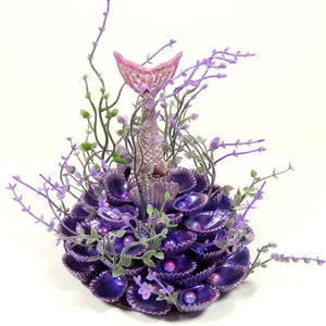 May include: A purple and pink mermaid tail figurine surrounded by purple seashells and greenery. The figurine is standing upright on a bed of seashells.