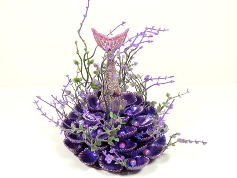 May include: A purple and pink mermaid tail figurine surrounded by purple seashells and greenery. The figurine is standing upright on a bed of seashells.