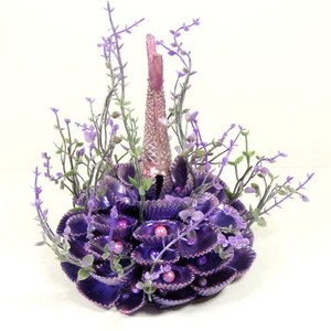 May include: A purple and pink mermaid tail made of seashells and faux flowers. The tail is surrounded by purple seashells and faux flowers.