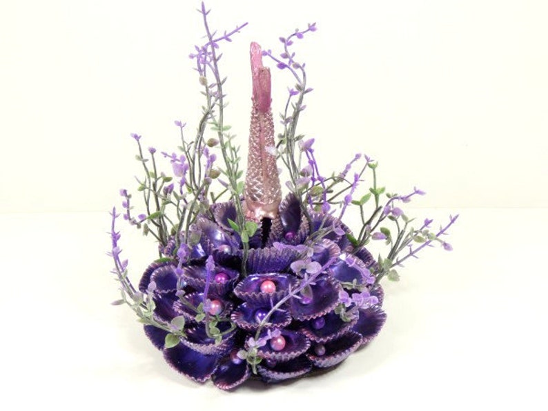 May include: A purple and pink mermaid tail made of seashells and faux flowers. The tail is surrounded by purple seashells and faux flowers.