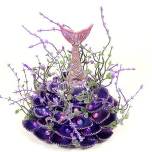 May include: A purple and pink mermaid tail figurine surrounded by purple seashells and lavender sprigs.
