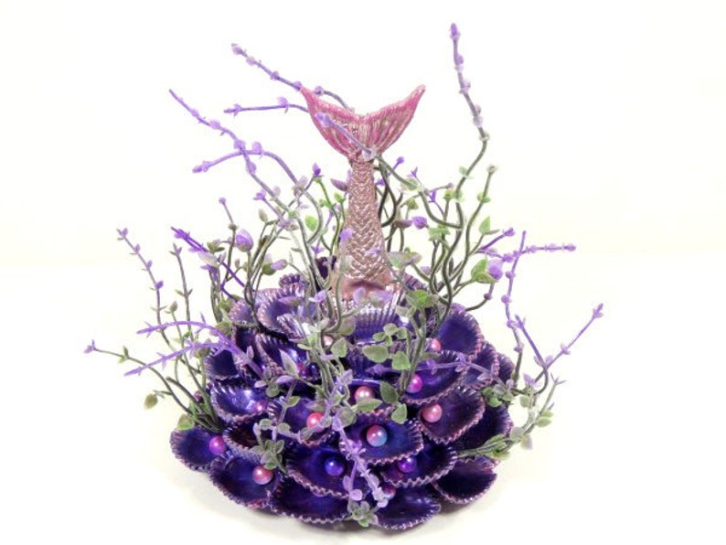 May include: A purple and pink mermaid tail figurine surrounded by purple seashells and lavender sprigs.