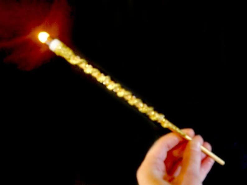 May include: A gold-coloured, twisted wand with a lit end. The wand is held in a person's hand.
