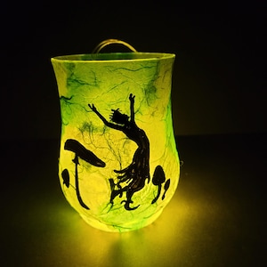 Könnte beinhalten: Eine leuchtend grüne Papierlaterne mit einer schwarzen Silhouette einer tanzenden Figur und Pilzen. Die Laterne hat einen gebogenen Griff und wird von innen beleuchtet, wodurch eine warme Atmosphäre entsteht. Es ist ein dekorativer Gegenstand.