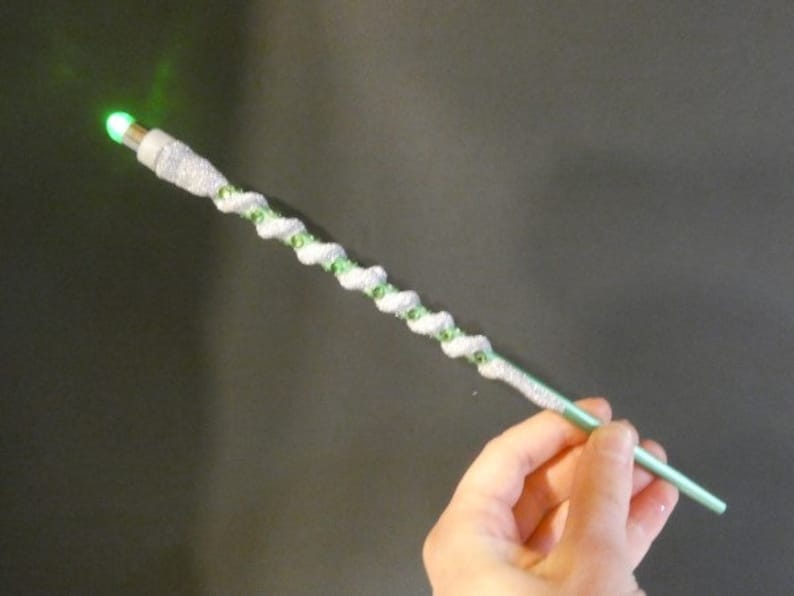 May include: A green and silver wand with a green light at the tip. The wand is decorated with silver glitter and green beads.