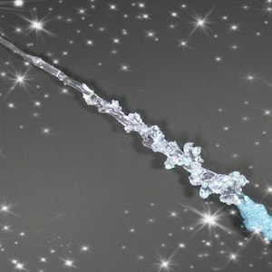 May include: A clear glass wand with a blue glitter tip. The wand is decorated with a frosted, icy design. The wand is set against a dark background with white stars.