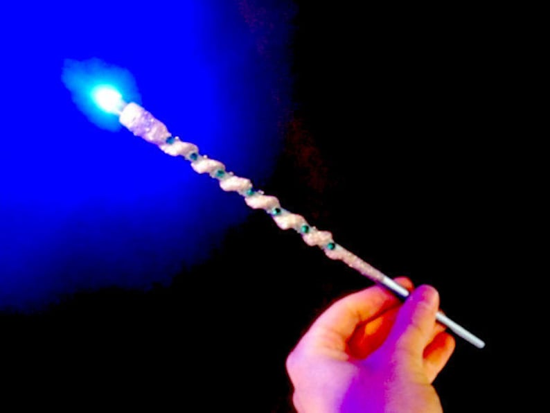 May include: A white wand with green glitter accents and a blue light at the tip. The wand is held in a hand.