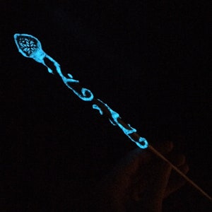 Può includere: Una bacchetta blu incandescente con disegni intricati. La bacchetta è fatta di legno e ha una punta appuntita. La bacchetta è tenuta in una mano, ma la mano non è visibile.