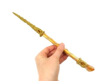 May include: A gold glitter wand with a clear crystal tip. The wand is held in a hand.