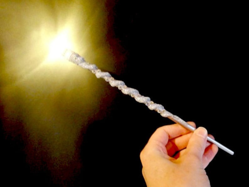 May include: A silver wand with a sparkly finish, casting a bright light. The wand is held in a hand against a black background.