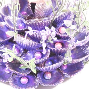 May include: A purple seashell arrangement with faux pearls and greenery. The seashells are arranged in a circular pattern and are accented with faux pearls and greenery.