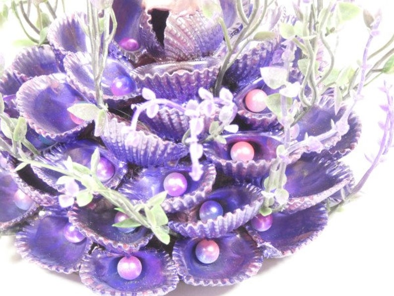 May include: A purple seashell arrangement with faux pearls and greenery. The seashells are arranged in a circular pattern and are accented with faux pearls and greenery.
