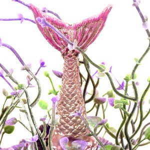 May include: A pink and gold mermaid tail figurine surrounded by purple flowers. The tail is detailed with scales and has a fin at the end.