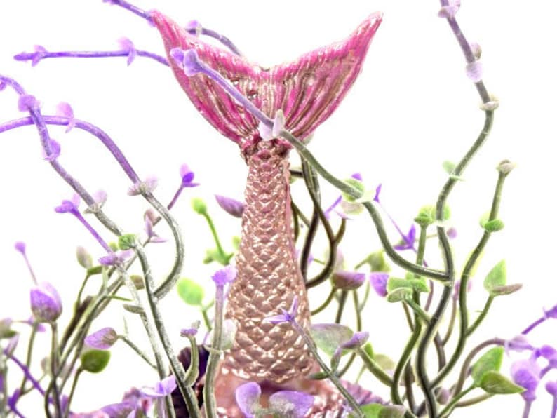 May include: A pink and gold mermaid tail figurine surrounded by purple flowers. The tail is detailed with scales and has a fin at the end.