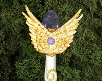 May include: A white and gold wand with a purple amethyst crystal at the top. The wand is decorated with gold wings and a smaller purple crystal.