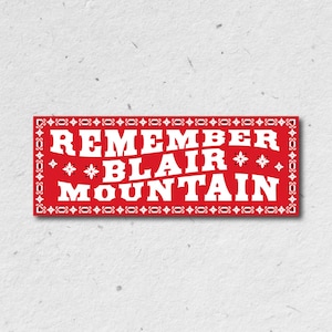 May include: A rectangular red sticker featuring the white text REMEMBER BLAIR MOUNTAIN in a bold, serif font. The design is enclosed by a white decorative border with repeating floral patterns and small star-like accents throughout the background.