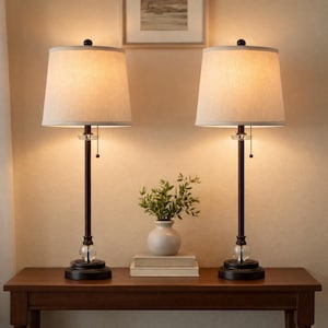 May include: Two matching table lamps with beige fabric shades and dark bronze bases. Each lamp has a clear crystal accent and a pull chain. The lamps are on a dark wood table, with a small vase of greenery and books.