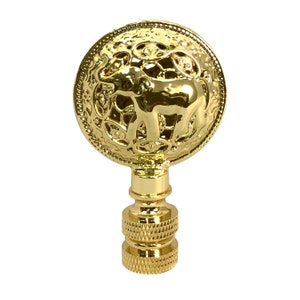 May include: Gold lamp finial with an ornate design featuring an elephant in the center. The finial is round and has a decorative pattern around the edge.