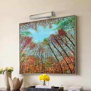 May include: A framed painting of a forest scene with tall trees and a blue sky. The trees are a mix of green, red, and yellow leaves. A silver light fixture is mounted above the painting.