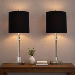 May include: Two matching table lamps with black shades and silver-colored bases. The lamps have a rectangular base and a crystal-like accent near the top of the stem. A small white vase with greenery and two books are on the table.