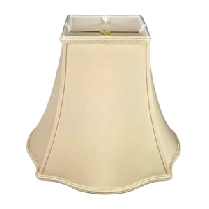 May include: A beige lampshade with a scalloped bottom edge and a decorative trim around the top. The shade is made of fabric and has a square shape at the top.