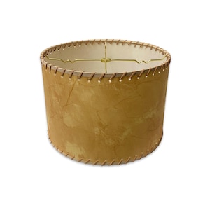 May include: A cylindrical lampshade with a textured, mottled tan exterior. The shade is stitched with a light brown thread along the top and bottom edges. A gold-colored metal frame supports the interior.