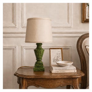 May include: A table lamp featuring a green marble-patterned base with a square pedestal and a tapered body, topped with a plain beige fabric lampshade. The lamp sits on a wooden surface next to two stacked books and a small white ceramic bowl.