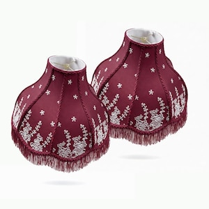 May include: Two bell-shaped lampshades in deep burgundy fabric featuring white embroidered floral and snowflake patterns. Each shade is accented with matching burgundy fringe trim along the scalloped bottom edge and white interior lining.