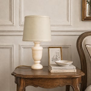May include: A cream-colored table lamp with a beige lampshade sits on a dark wooden table. The lamp has a marble-like base. Beside the lamp are books, a small bowl, and a framed picture. The table is in front of a wall with paneling.